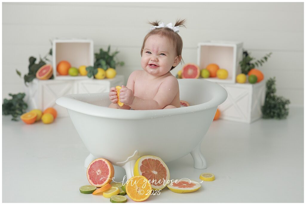 One year old girl sitting in bathtub for citrus fruit milk bath first birthday photos, making sour faces because she is eating the fruit