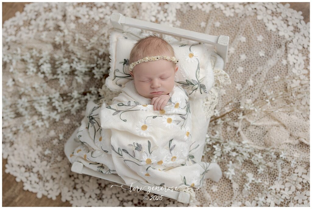 Beautiful newborn baby girl photographed by Lehigh Valley newborn photographer Lori Generose in Lehigh Valley, PA – posed in a cozy wooden newborn bed with lace blanket underneath and a boho daisy wrap and pillow during a professional studio newborn session.