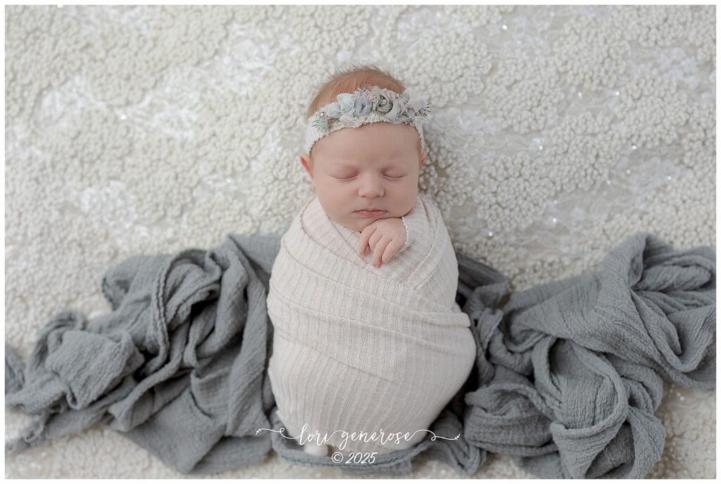 Beautiful newborn baby girl photographed by Lehigh Valley newborn photographer Lori Generose in Lehigh Valley, PA – wrapped in a white wrap on a lace blanket with a gray wrap splayed out under her and floral headband during a professional studio newborn session.