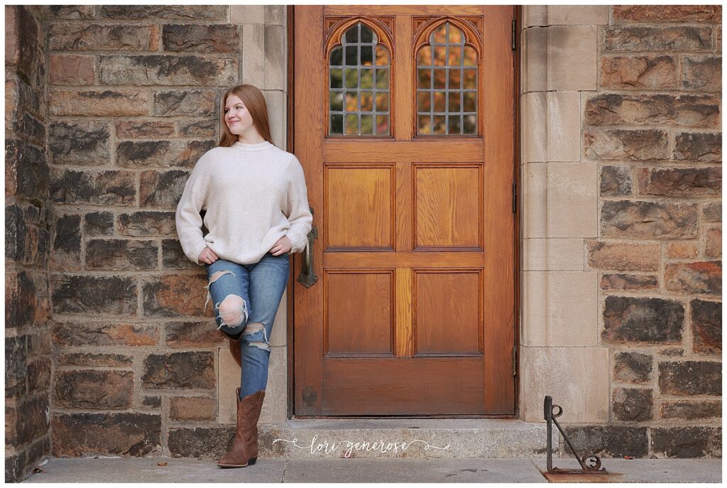 Senior girl poses confidently in front of a historic wooden door at Lehigh University in Bethlehem, PA during a fall senior session with Lehigh Valley photographer Lori Generose. Her outfit—an oversized neutral sweater, ripped jeans, and cowboy boots offers a cozy, casual style that’s perfect for senior portraits with a rustic, collegiate vibe.