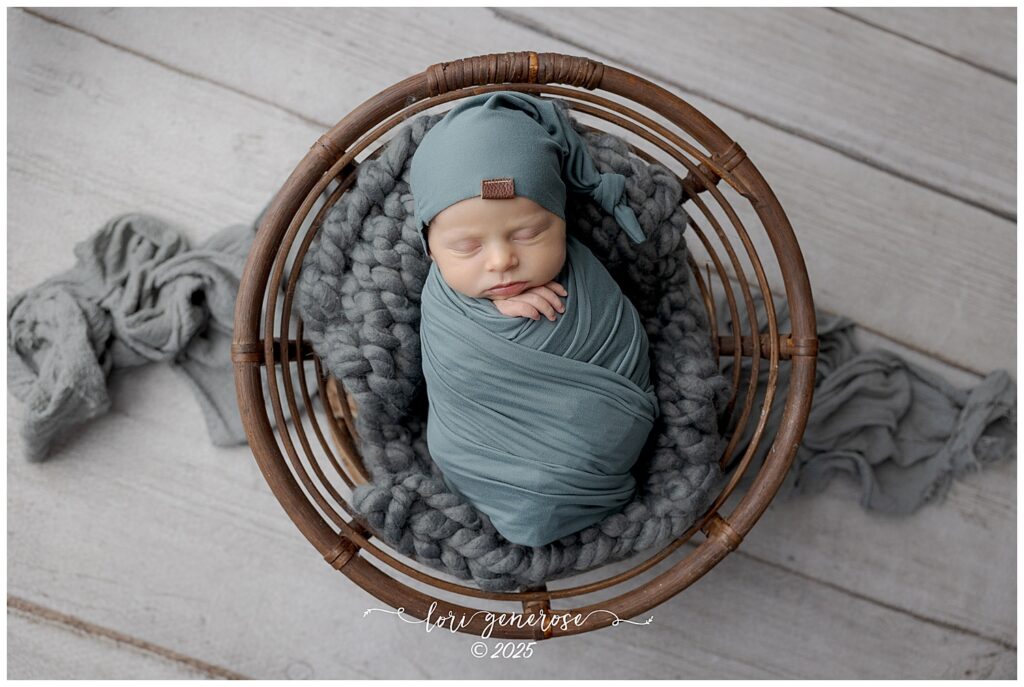 Newborn baby boy sleeping swaddled in a soft dusty blue wrap with a matching sleepy cap, posed in a round wicker basket lined with a chunky gray knit blanket on a light wooden floor. Studio newborn portrait by LG Photography- Lori Generose, Lehigh Valley PA newborn photographer.