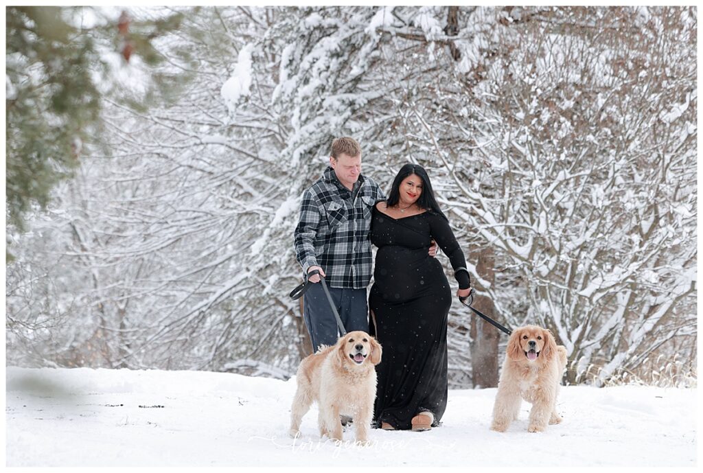Maternity portrait by Lori Generose of LG Photography, a Lehigh Valley maternity and newborn photographer, featuring an expecting couple walking outdoors in a snow-covered landscape, with mom in a black floor-length maternity dress, dad in a blue plaid flannel and jeans and their dogs (golden rertievers mixed with cocker spaniels) surrounded by snow-covered trees.