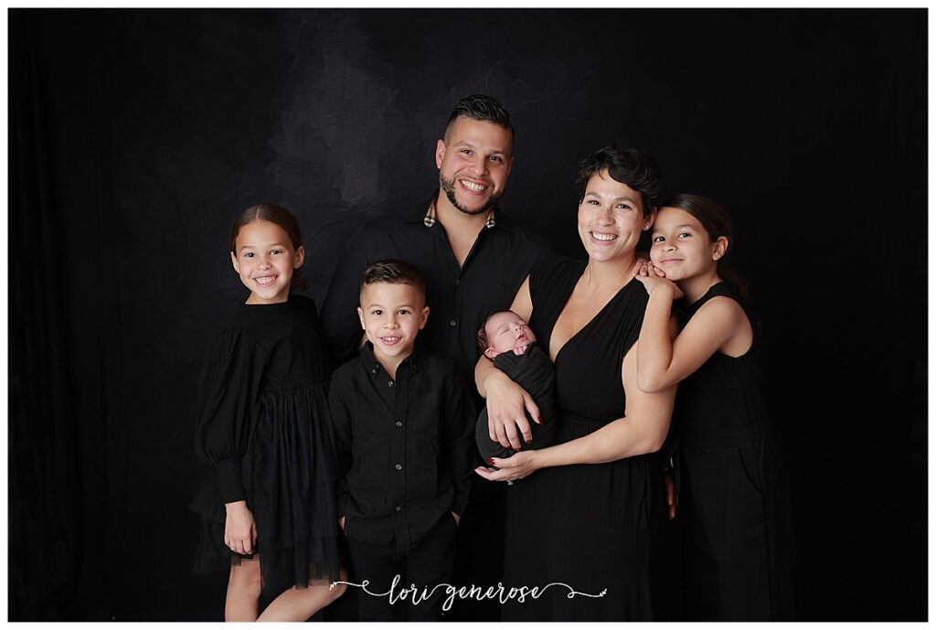 Newborn family portrait by LG Photography, a Lehigh Valley newborn photographer, featuring parents and siblings styled in classic black outfits against a dark studio backdrop with a wrapped newborn- elegant inspiration for what to wear for your newborn photography session.