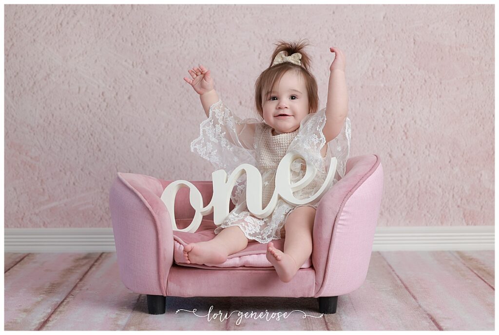 First birthday portrait session for baby girl with blush pink backdrop sitting on a small pink couch with a cursive one sign on her lap with her hands up from Lori Generose, who is a LG Photography- a Lehigh Valley cake smash photographer