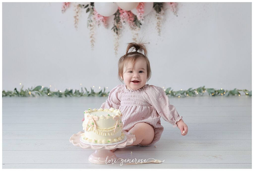 One year old girl cake smash portraits with pink and cream balloon garland with pink hanging wisteria flowers. Baby has a cream cake with vintage and pink bows from LG Photography, Lori Generose who is a Lehigh Valley cake smash photographer
