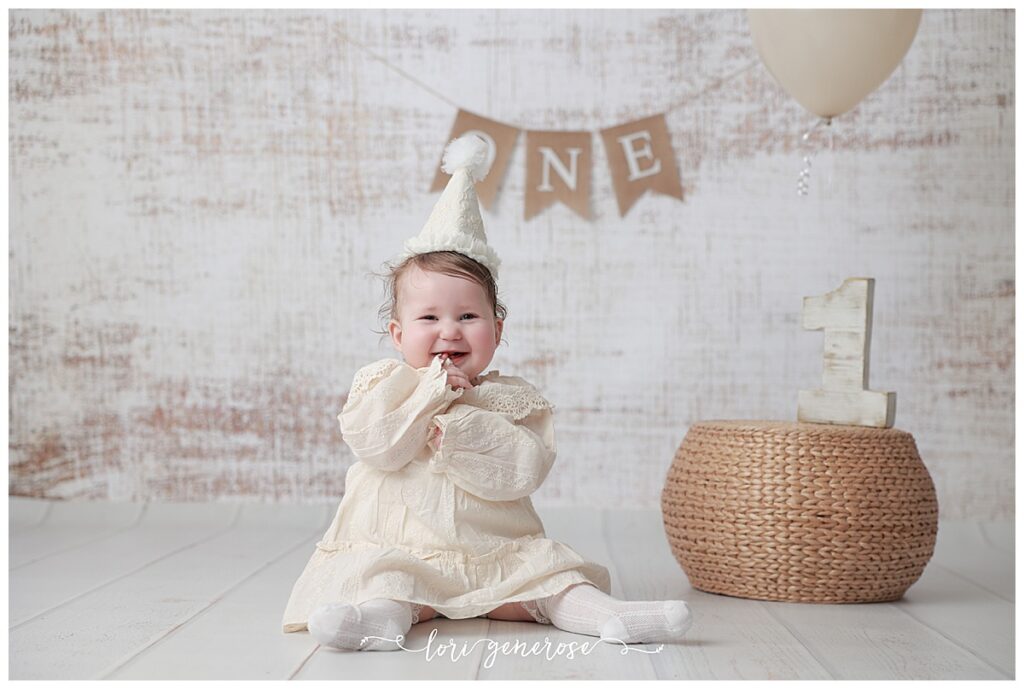 First birthday girl in a cream classic dress with a cream cone party hat for her cake smash session with Lori Generose of LG Photography, a Lehigh Valley Photographer