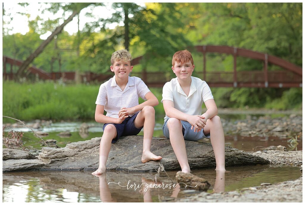 Family photoshoot of mom, dad and their two boys ages 11 and 13 in the water posing on a rock with a brown iron bridge behind them at Trexler Nature Preserve in Schnecksville, PA by Lehigh Valley Photographer, Lori Generose of LG Photography