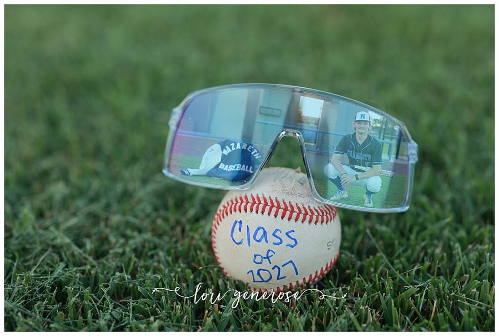 senior boy photoshoot with a photo of his sunglasses with a reflection of the Nazareth High School Baseball senior reflected in them and his letterman Nazareth Baseball jacket and a baseball with Class of 2027 written on it at a baseball field in Nazareth, PA all taken by Lehigh Valley Senior photographer Lori Generose of LG Photography